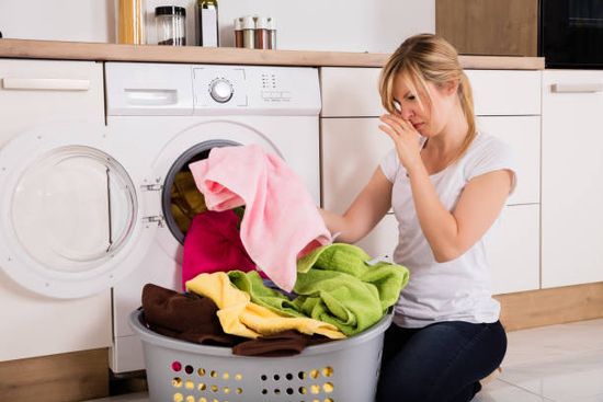 Young Woman Looking At Smelly Clothes Out Of Washing Machine In Kitchen